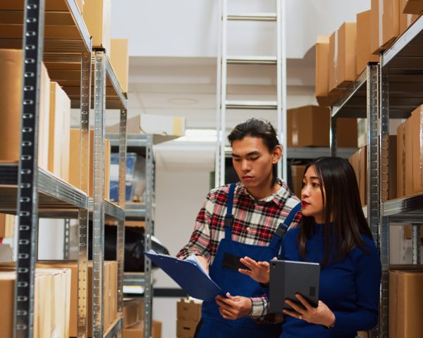 Asian employees checking warehouse supplies on tablet and files, working with storage room logistics. Young team of people using gadget and papers for business plan and production.